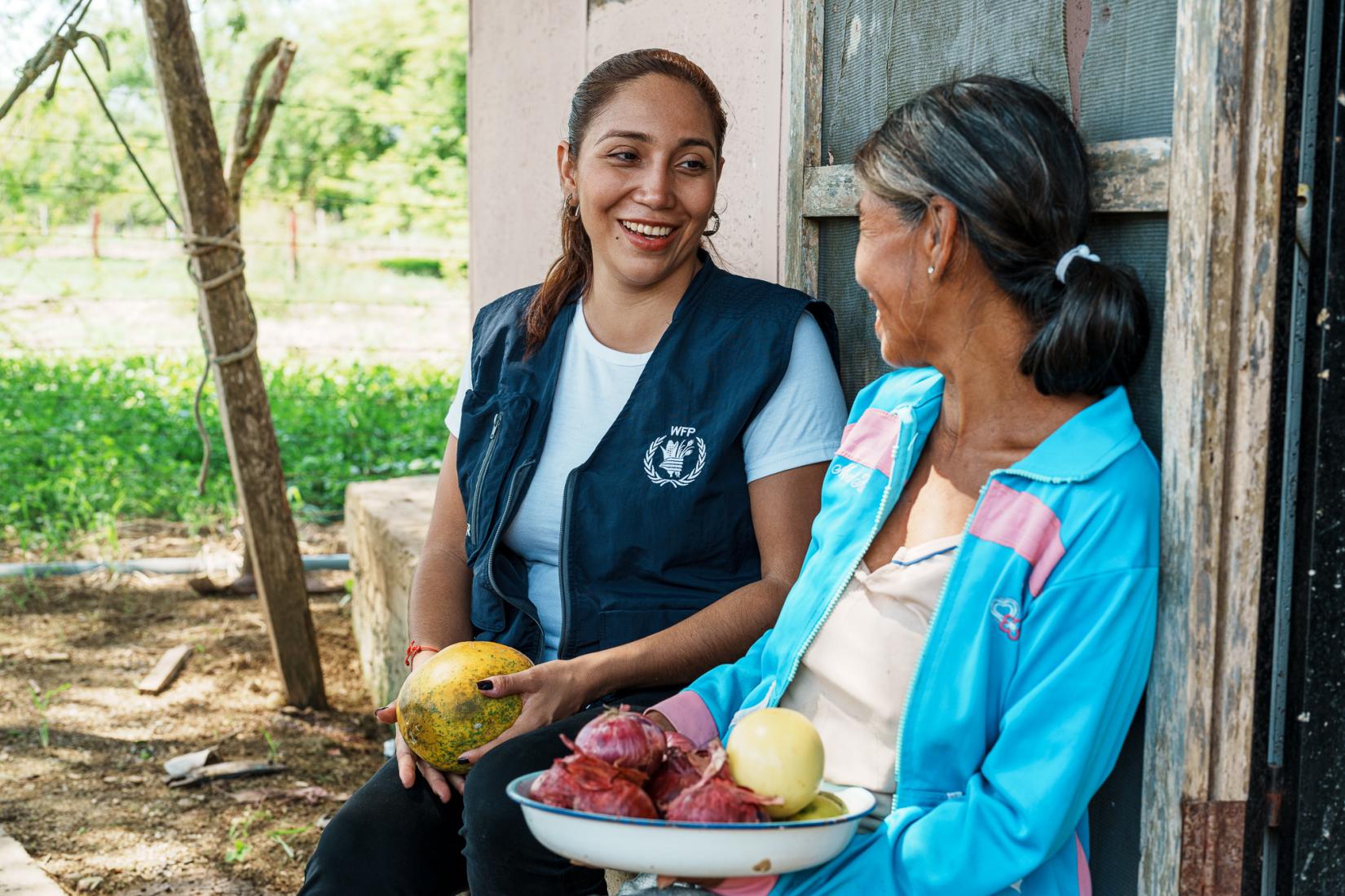 Trabajadora del Programa Mundial de Alimentos entregando comida como parte del proyecto de comidas escolares a una mujer que parece pertenecer a la institución. Ambas se encuentran sentadas, una la mira a la otra con una sonrisa y un bowl de frutas en sus manos.